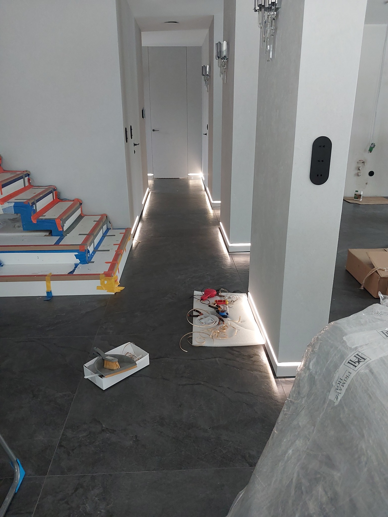 Korytarz w trakcie wykańczania z listwami przypodłogowymi LED, schody zabezpieczone taśmą malarską, widoczne narzędzia i opakowania po materiałach budowlanych.
