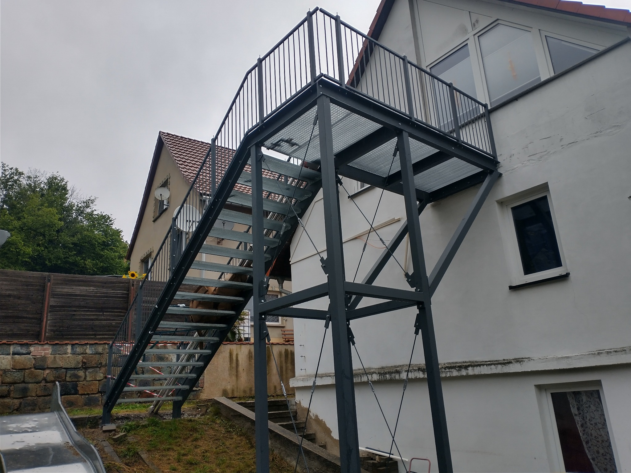 Zewnętrzne metalowe schody z platformą i balustradą, prowadzące do górnego piętra budynku, widoczne elementy mocujące i drabinka dla dzieci w tle.