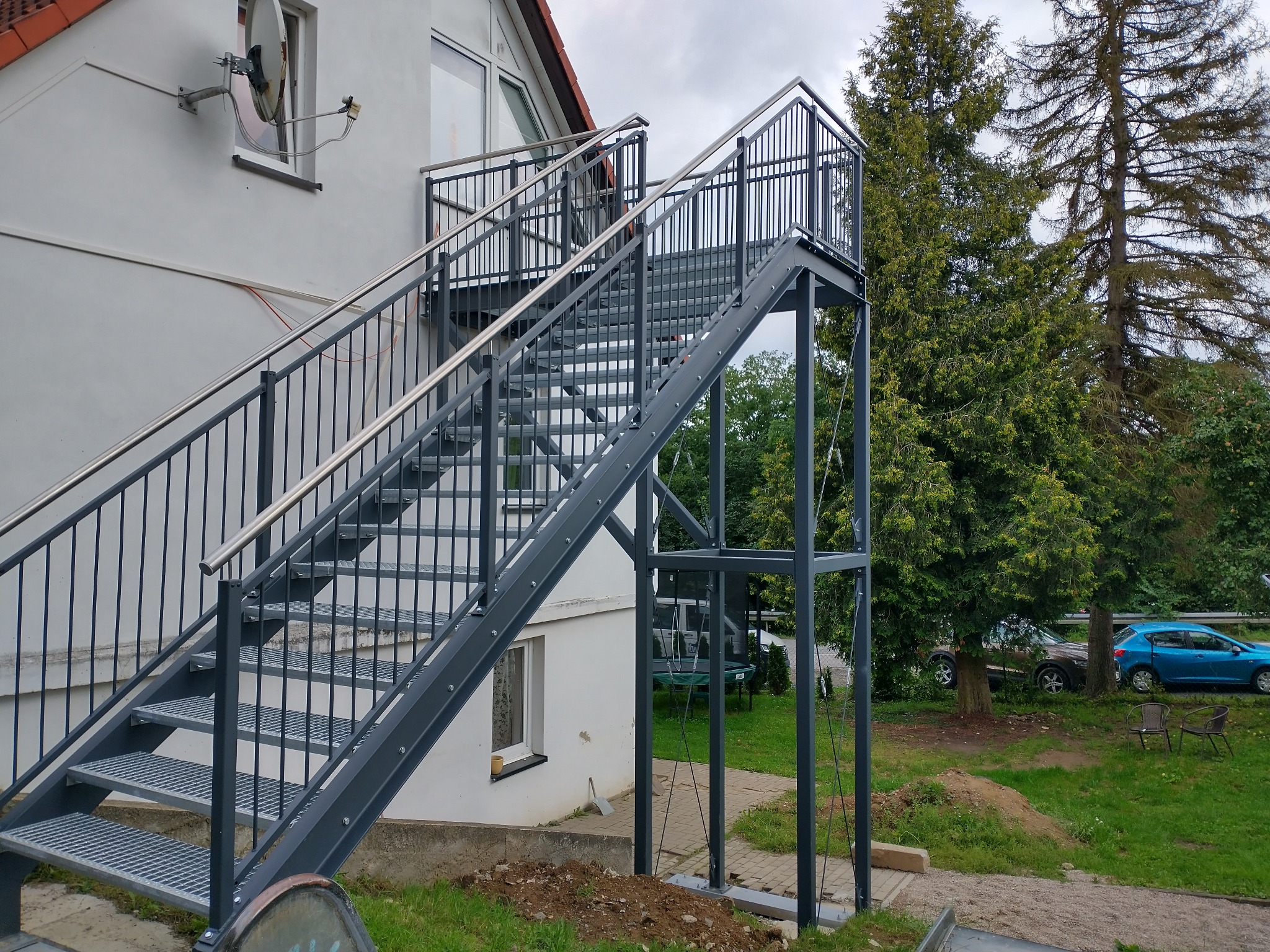 Zewnętrzne schody metalowe z balustradą ze stali nierdzewnej prowadzące do wejścia na piętro budynku mieszkalnego, widoczne drzewa i samochód w tle.