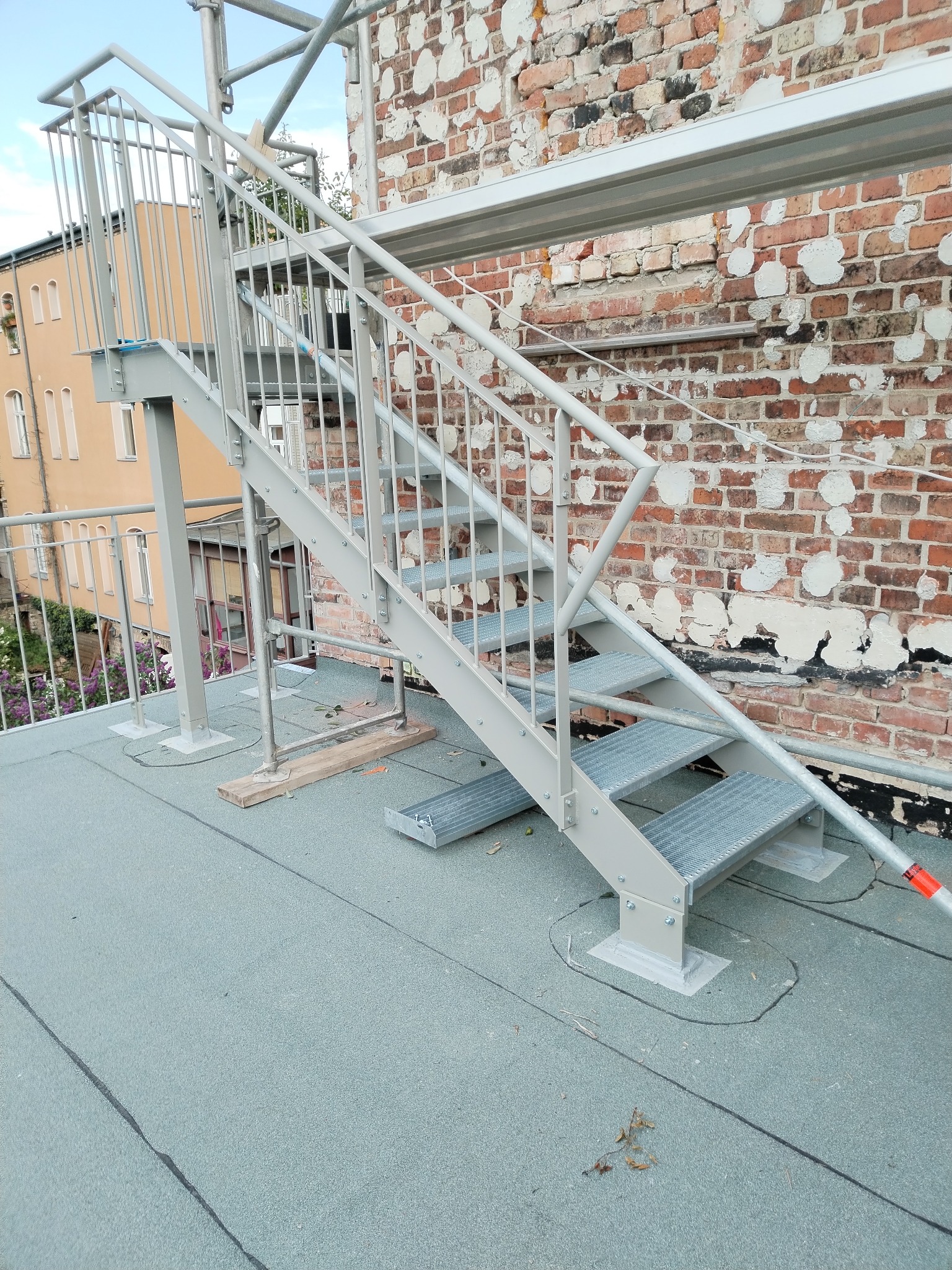 Szare metalowe schody zewnętrzne z kratowanymi stopniami, zamontowane na dachu z papy, z ceglaną ścianą w tle.