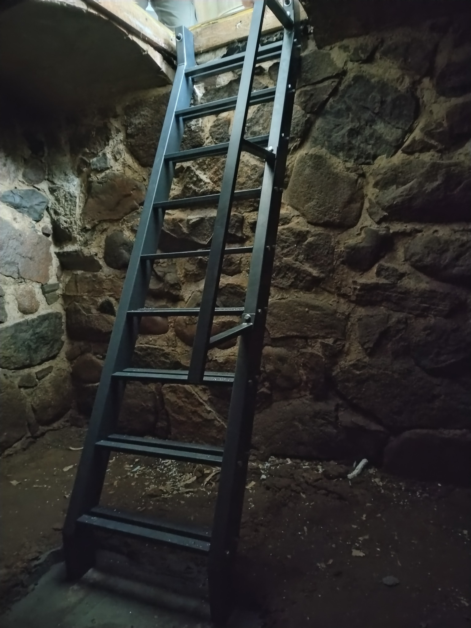 Stalowa drabina przymocowana do kamiennej ściany, prowadząca do otworu w suficie z widocznymi elementami drewnianej konstrukcji.