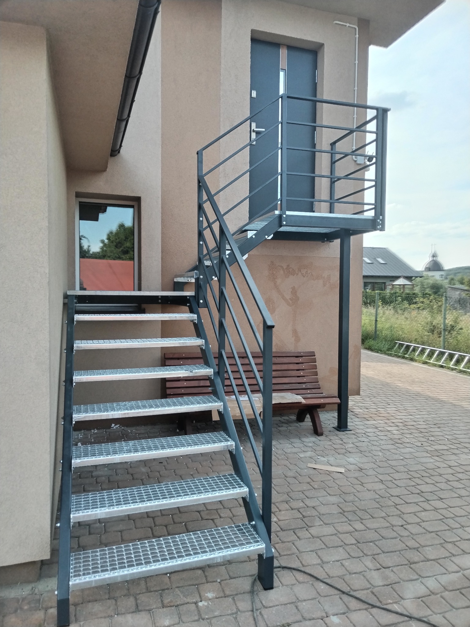 Zewnętrzne schody z metalową konstrukcją prowadzące do drzwi na podwyższeniu, z metalową balustradą, na tle budynku z ceglaną ścieżką i ławką.