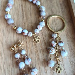 Beztroski Świat - Komplet biżuterii: bransoletka z koralików, kolczyki sztyfty i brelok z zawieszką w kształcie czterolistnej koniczyny, wszystko w tonacji bieli, brązu i złota, na drewnianej tacy.