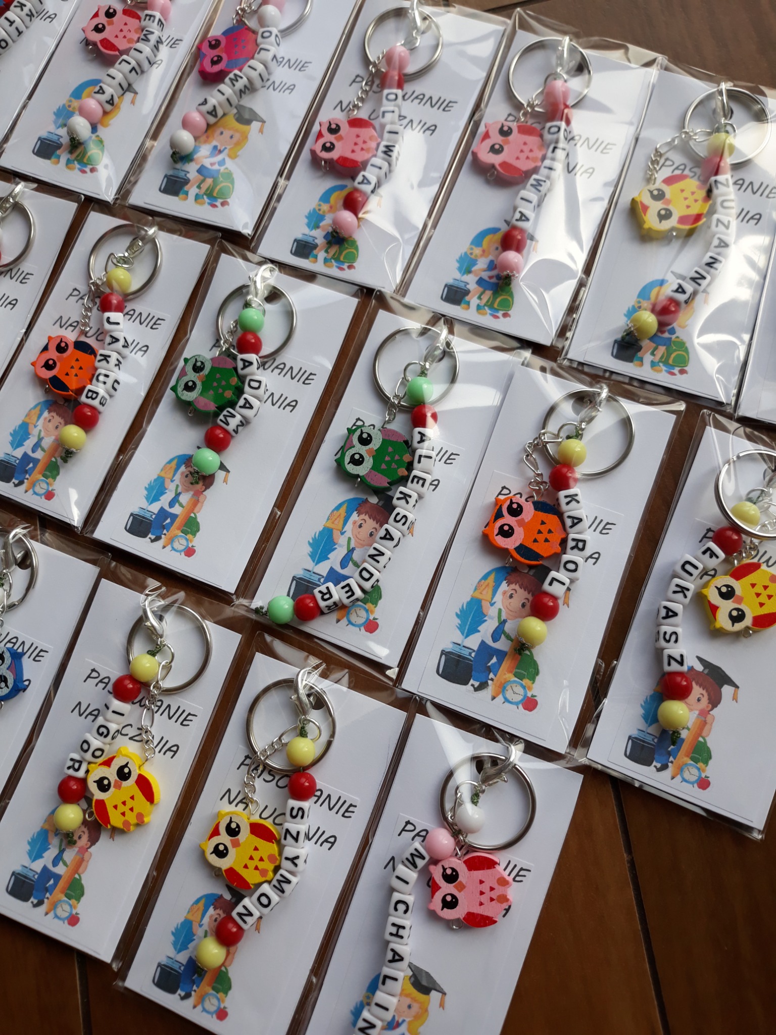 Ręcznie robione breloczki z imionami, zawieszkami w kształcie sowy i kolorowymi koralikami, zapakowane w foliowe woreczki z kartonikami z motywem ucznia.