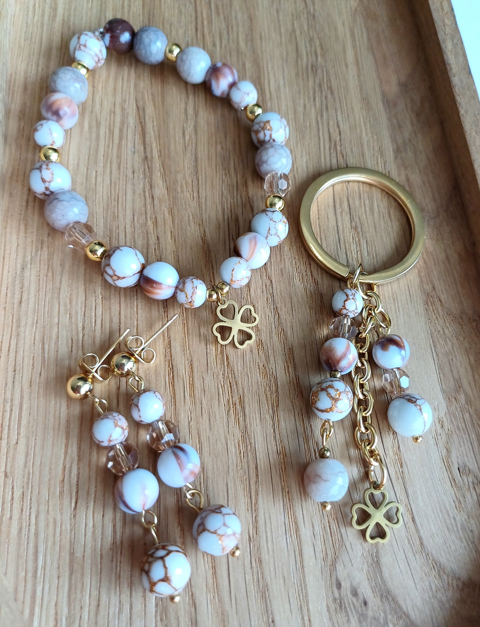 Komplet biżuterii: bransoletka z koralików, kolczyki sztyfty i brelok z zawieszką w kształcie czterolistnej koniczyny, wszystko w tonacji bieli, brązu i złota, na drewnianej tacy.