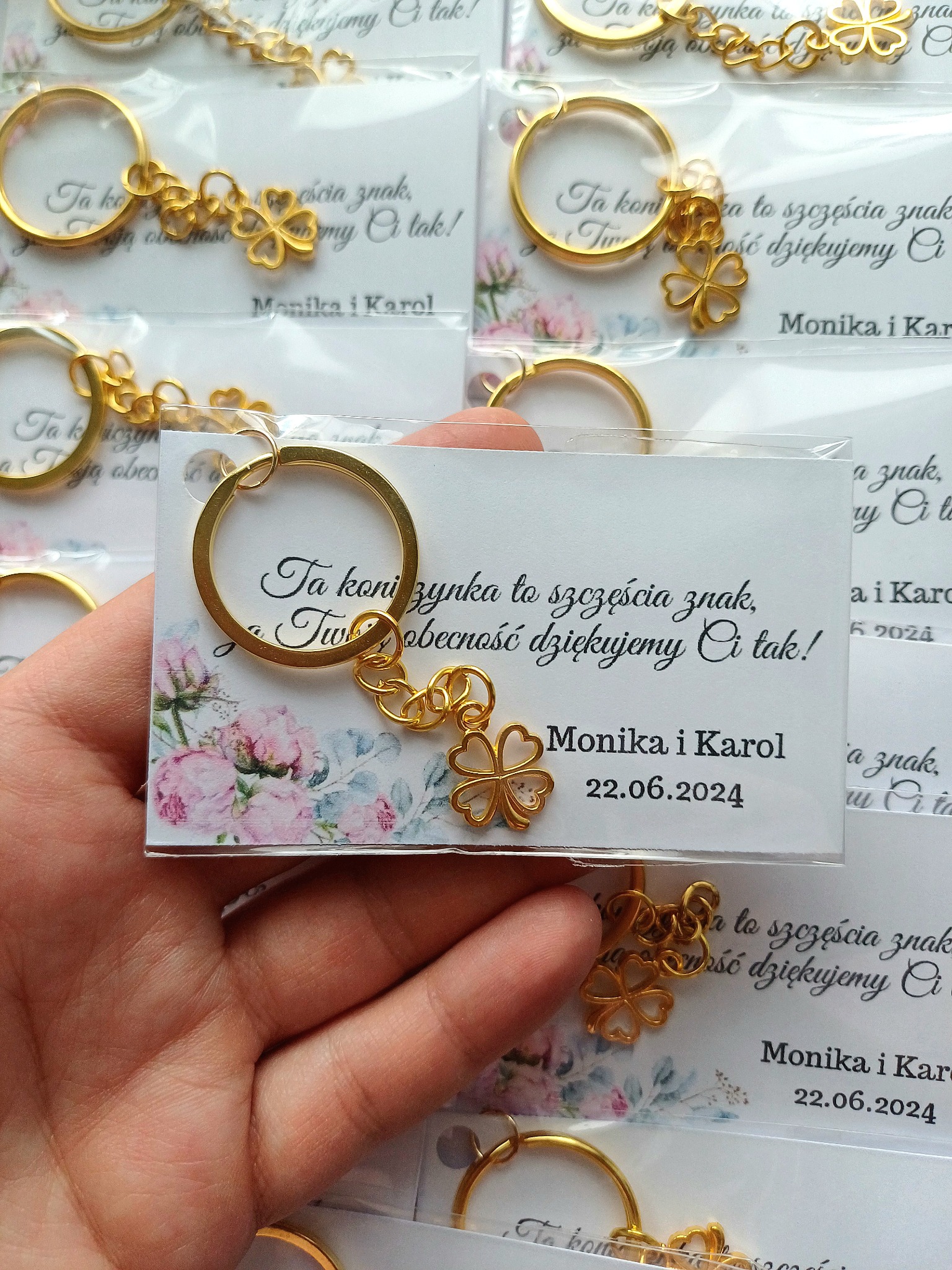 Złoty breloczek z czterolistną koniczyną, przymocowany do kartonika z podziękowaniem dla gości weselnych od Moniki i Karola, data ślubu 22.06.2024, prezentowany na dłoni na tle innych, podobnych...
