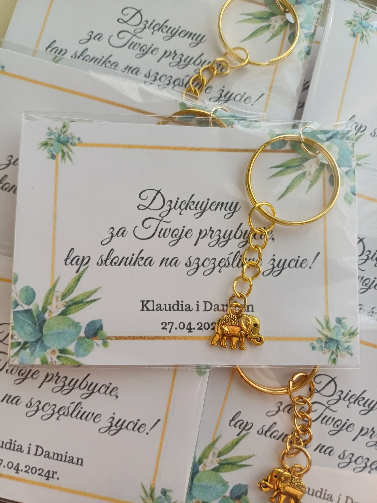 Podziękowania dla gości w Ostrołęce: złoty breloczek-słonik z kartką 'Dziękujemy za Twoje przybycie, łap słonika na szczęśliwe życie!' zapakowany w folię.