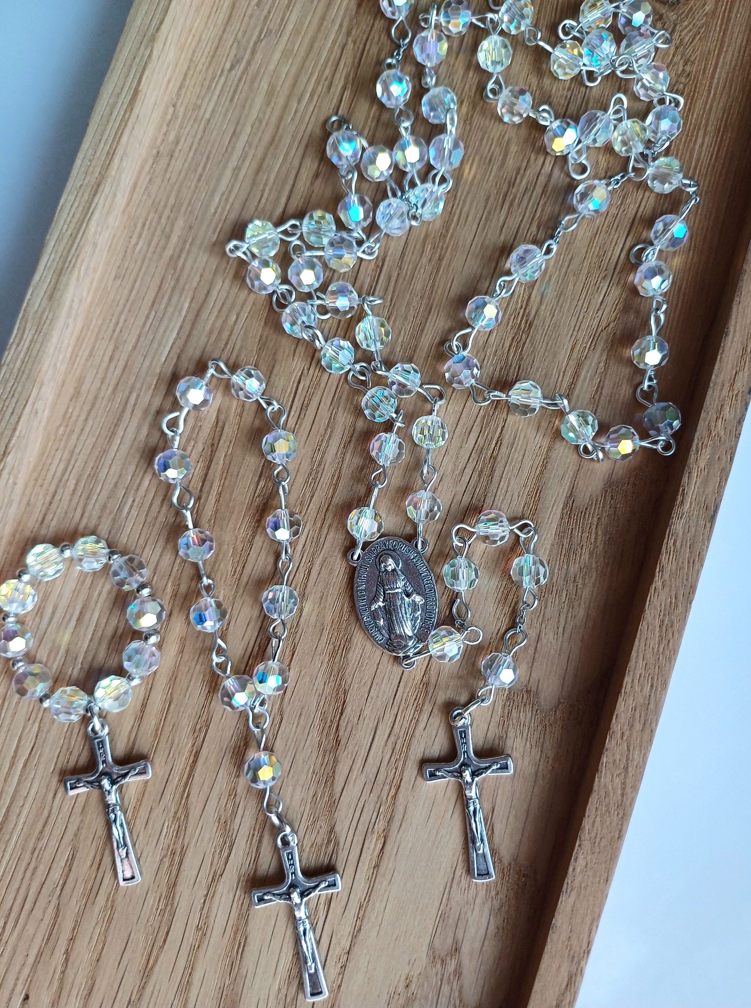 Trzy różańce z błyszczących, fasetowanych koralików i srebrnymi krzyżykami na drewnianej tacy, jeden z medalikiem Matki Bożej.