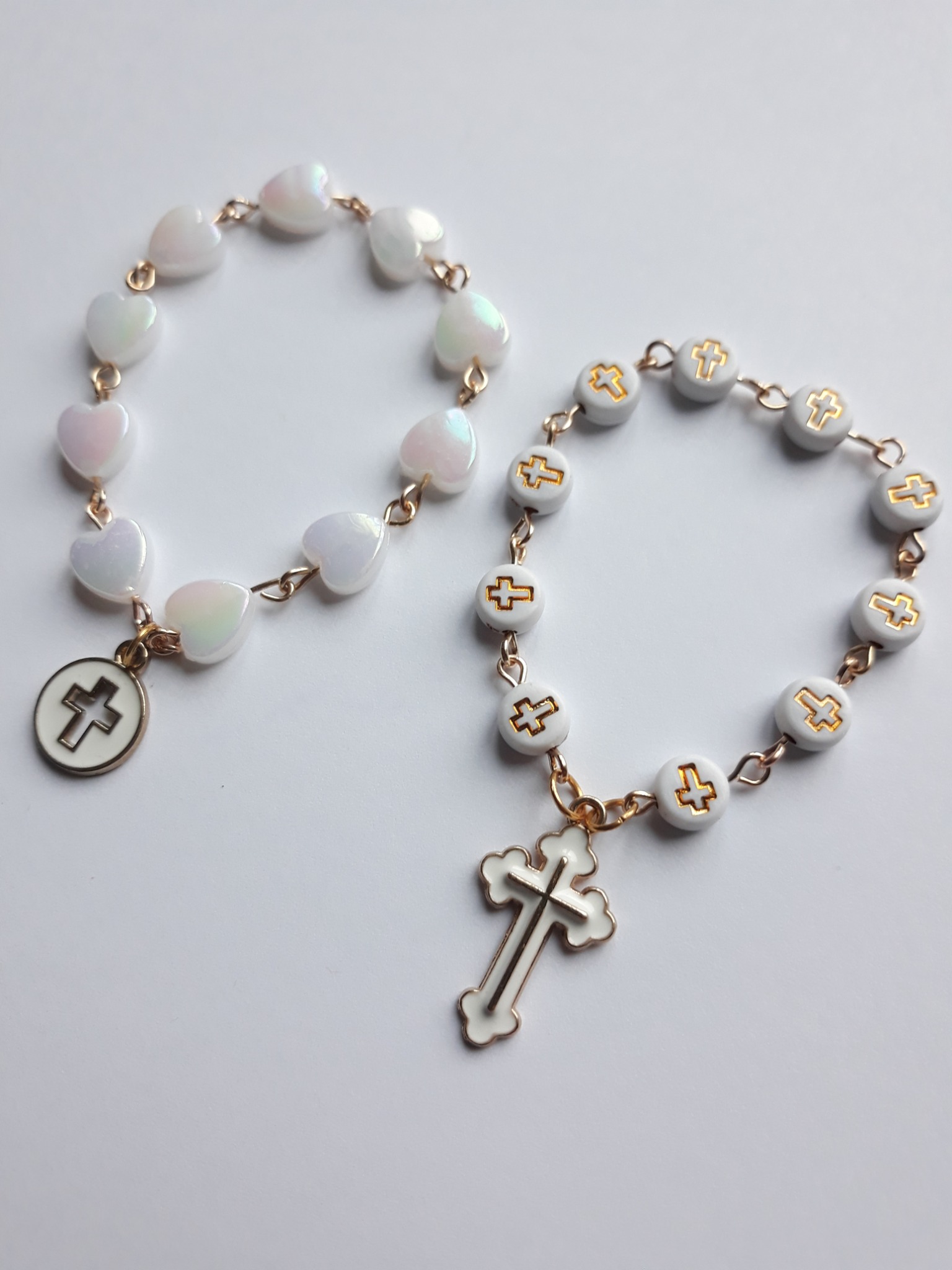 Dwa różańce: jeden z białych koralików w kształcie serc z opalizującym połyskiem i zawieszką z krzyżem w okręgu, drugi z białych okrągłych koralików z złotymi krzyżami i zawieszką z krzyżem.