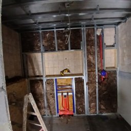 Usługi remontowo budowlane BUDSOB - Wnętrze w trakcie remontu: metalowy szkielet ścianki działowej wypełniony wełną mineralną, instalacja hydrauliczna w kolorze czerwonym i niebieskim, drabina oparta o ścianę z widocznymi śladami...