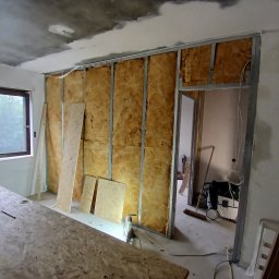 Usługi remontowo budowlane BUDSOB - Montaż ścianki działowej: metalowy szkielet z izolacją z wełny mineralnej, płyty OSB oparte o ścianę, surowy betonowy sufit w tle, widoczny odkurzacz przemysłowy.