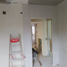 Usługi remontowo budowlane BUDSOB - Montaż płyt kartonowo-gipsowych wokół otworu drzwiowego w trakcie remontu. Widoczna drabina, odkurzacz przemysłowy i instalacja elektryczna.