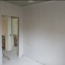 Wnętrze w trakcie montażu płyt kartonowo-gipsowych, widoczne łączenia płyt i wkręty, fragment drabiny malarskiej po lewej, widok na sąsiednie pomieszczenie przez otwór drzwiowy.