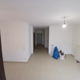 Usługi remontowo budowlane BUDSOB - Wnętrze pustego, jasnego mieszkania po remoncie. Widoczne białe ściany, beżowa podłoga, narzędzia malarskie: wiadro z farbą, pędzle, wałek oraz niebieska folia ochronna.