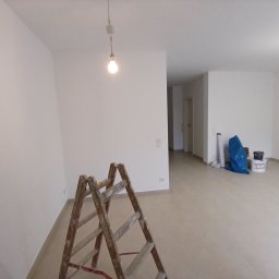 Usługi remontowo budowlane BUDSOB - Jasne, przestronne wnętrze w trakcie odświeżania, z drewnianą drabiną malarską na pierwszym planie, białe ściany po malowaniu, widoczne akcesoria malarskie w tle, surowa żarówka zwisająca z sufitu.