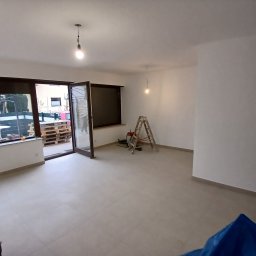 Usługi remontowo budowlane BUDSOB - Wnętrze świeżo wyremontowanego pokoju z szarymi płytkami podłogowymi, widoczne otwarte okno balkonowe, drabina malarska i wiadro z mopem.