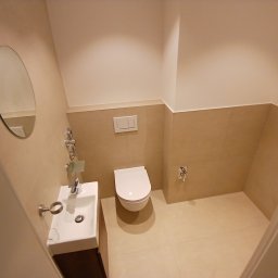 Usługi remontowo budowlane BUDSOB - Nowoczesna łazienka z beżowymi płytkami, owalnym lustrem, podwieszaną toaletą i umywalką z ciemnym drewnianym blatem, widziana z perspektywy wejścia.