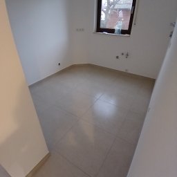 Usługi remontowo budowlane BUDSOB - Puste pomieszczenie z beżowymi płytkami podłogowymi, świeżo pomalowanymi białymi ścianami i oknem z widokiem na drzewo z karmnikiem dla ptaków, butelka wody na parapecie.