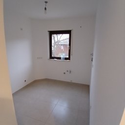Usługi remontowo budowlane BUDSOB - Puste pomieszczenie po remoncie z szarymi płytkami podłogowymi, świeżo pomalowanymi białymi ścianami, oknem z widokiem na podwórze i wiszącą żarówką bez klosza.