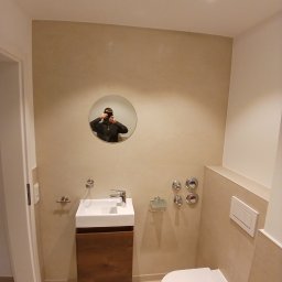 Usługi remontowo budowlane BUDSOB - Nowoczesna łazienka z beżowymi ścianami, wiszącą szafką z umywalką z drewna, toaletą i okrągłym lustrem, w którym odbija się osoba robiąca zdjęcie.