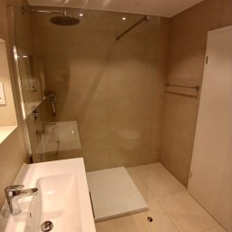 Usługi remontowo budowlane BUDSOB - Nowoczesna łazienka z beżowymi płytkami, szklaną kabiną prysznicową typu walk-in z deszczownicą, białą umywalką z chromowaną baterią i punktowym oświetleniem sufitowym, widoczny fragment odpływu...
