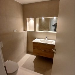 Usługi remontowo budowlane BUDSOB - Nowoczesna łazienka z beżowymi płytkami, drewnianą szafką pod umywalką, lustrem z oświetleniem i toaletą. Widoczne wnęki w ścianie z dodatkowym oświetleniem.