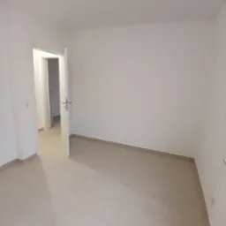 Pusta, jasna przestrzeń z beżową terakotą na podłodze, białe ściany i otwarte drzwi do kolejnego pomieszczenia, widoczne gniazdko elektryczne po lewej stronie.