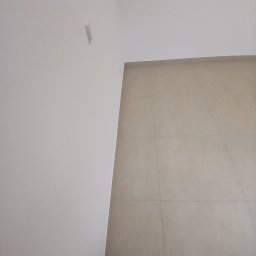 Usługi remontowo budowlane BUDSOB - Świeżo położone, beżowe płytki podłogowe w rogu pomieszczenia, widoczna biała ściana z niedużym śladem po usuniętym elemencie oraz fragment buta w dolnym rogu.