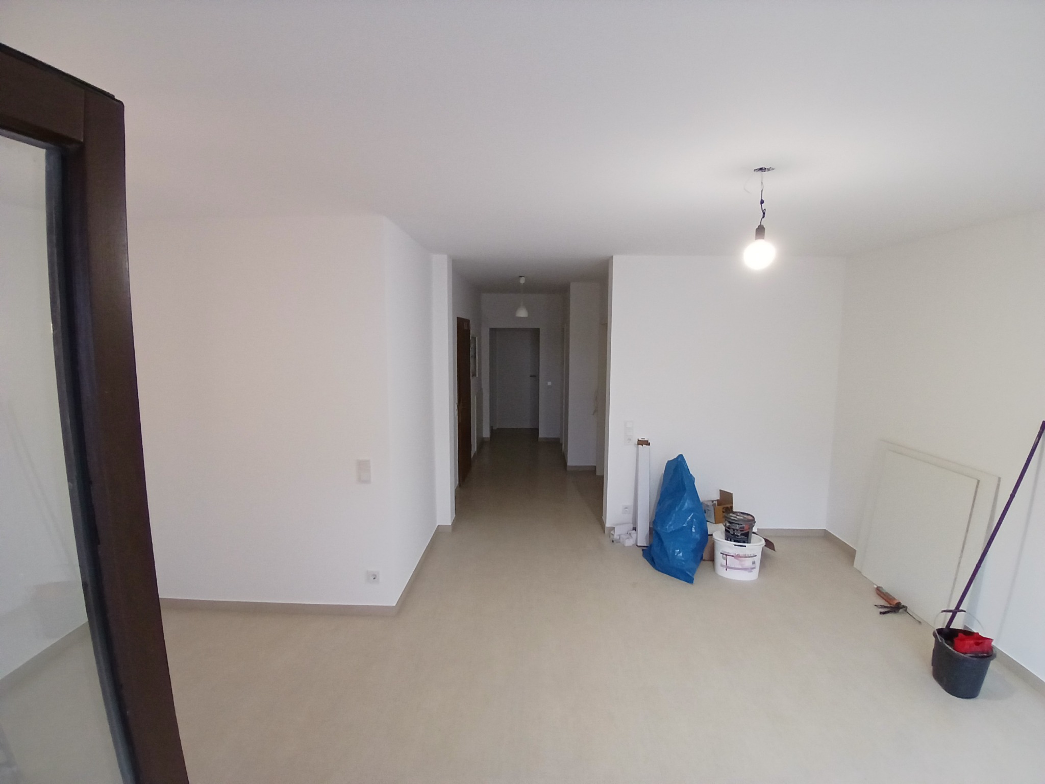 Wnętrze pustego, jasnego mieszkania po remoncie. Widoczne białe ściany, beżowa podłoga, narzędzia malarskie: wiadro z farbą, pędzle, wałek oraz niebieska folia ochronna.