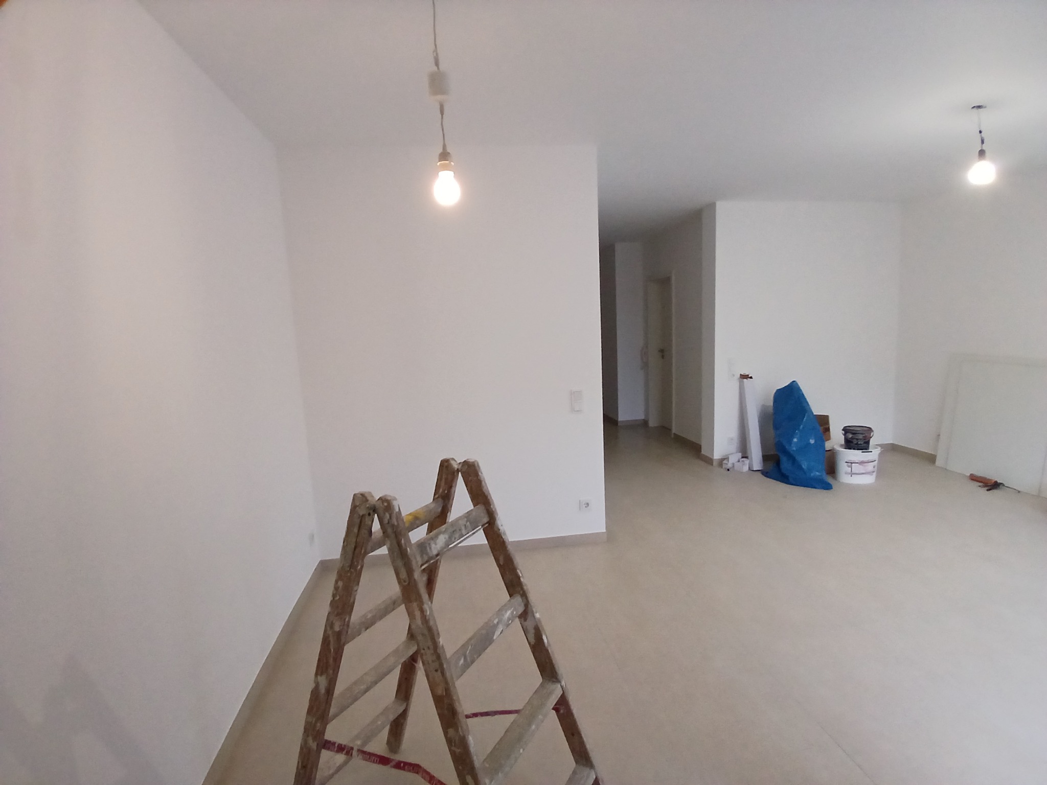 Jasne, przestronne wnętrze w trakcie odświeżania, z drewnianą drabiną malarską na pierwszym planie, białe ściany po malowaniu, widoczne akcesoria malarskie w tle, surowa żarówka zwisająca z sufitu.