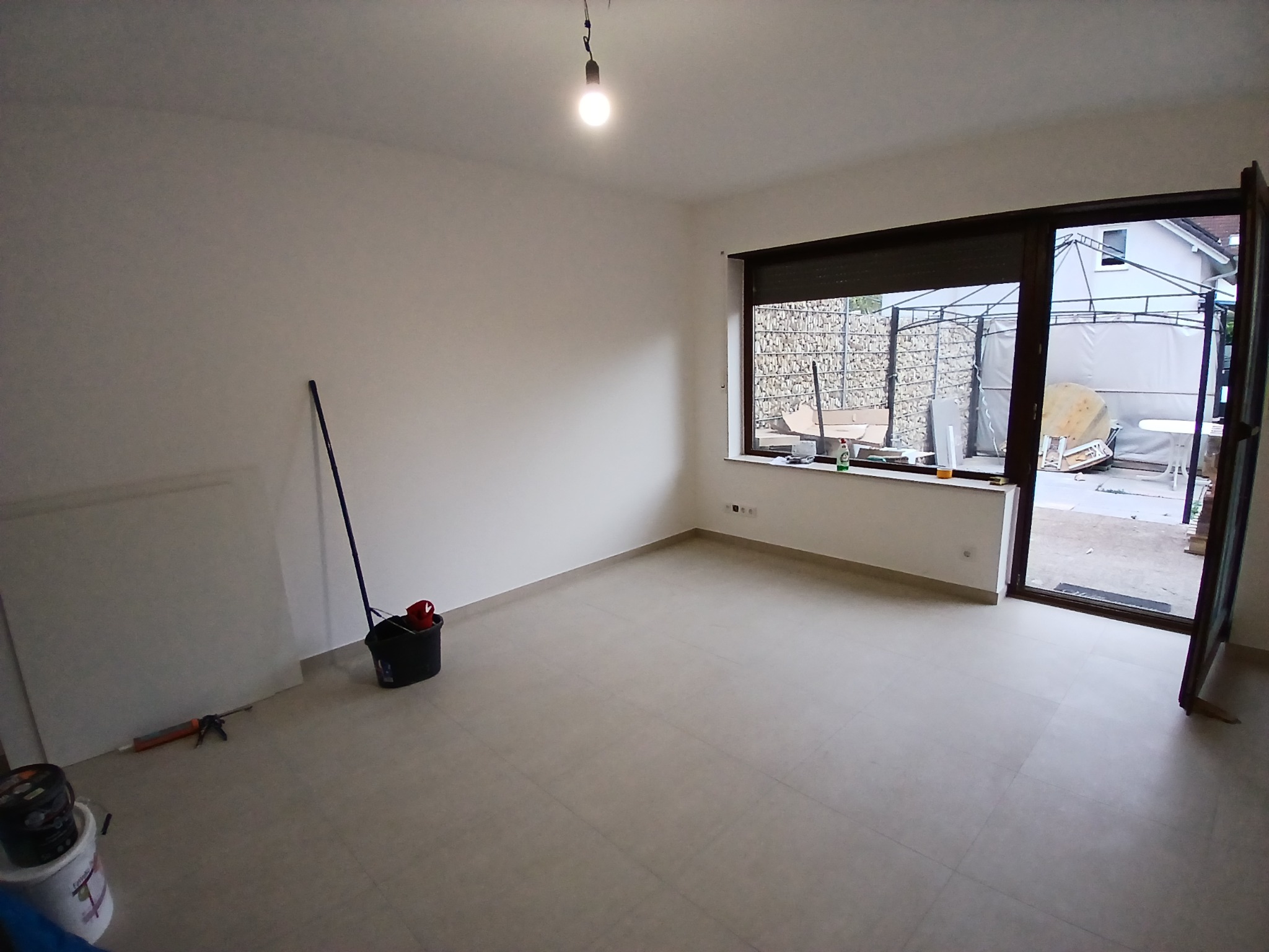 Przygotowane do malowania, puste pomieszczenie z białymi ścianami, widoczne narzędzia malarskie: wiadro, wałek, pistolet do silikonu, niezabezpieczona podłoga z płytek