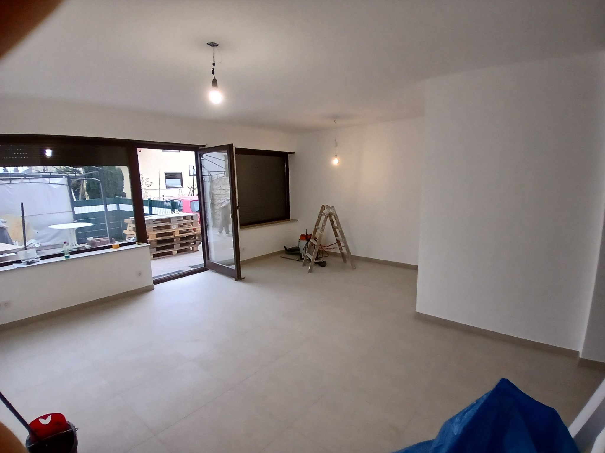 Wnętrze świeżo wyremontowanego pokoju z szarymi płytkami podłogowymi, widoczne otwarte okno balkonowe, drabina malarska i wiadro z mopem.