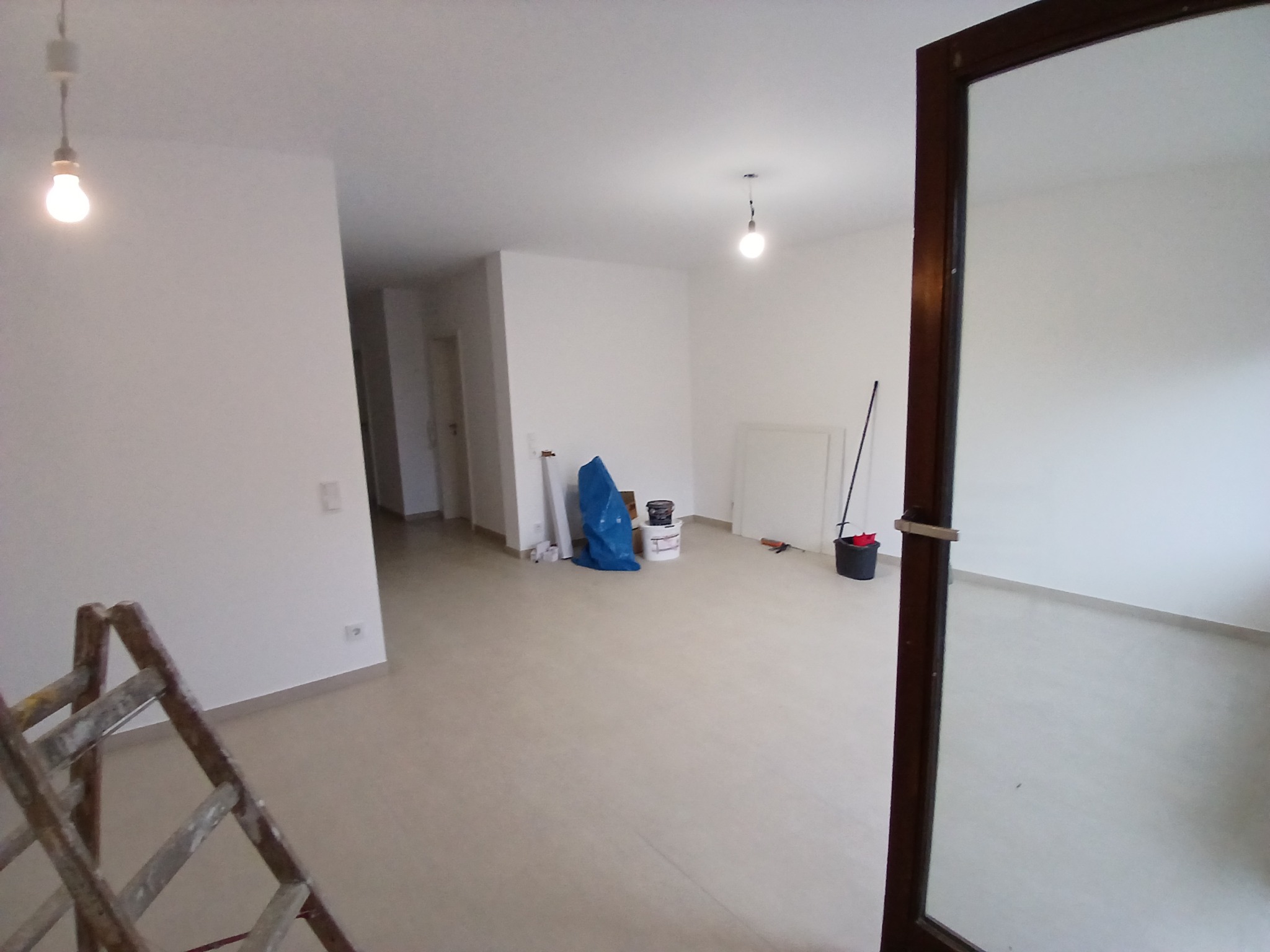Wnętrze w trakcie prac wykończeniowych: białe ściany, jasna podłoga, drabina malarska w rogu, wiadro z farbą i folia malarska w tle, widoczne żarówki sufitowe.