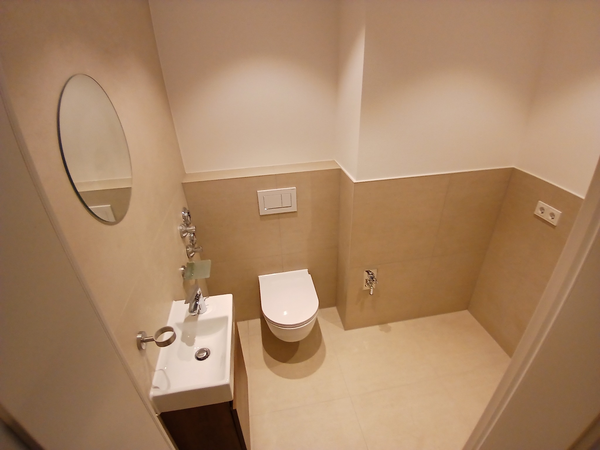 Nowoczesna łazienka z beżowymi płytkami, owalnym lustrem, podwieszaną toaletą i umywalką z ciemnym drewnianym blatem, widziana z perspektywy wejścia.