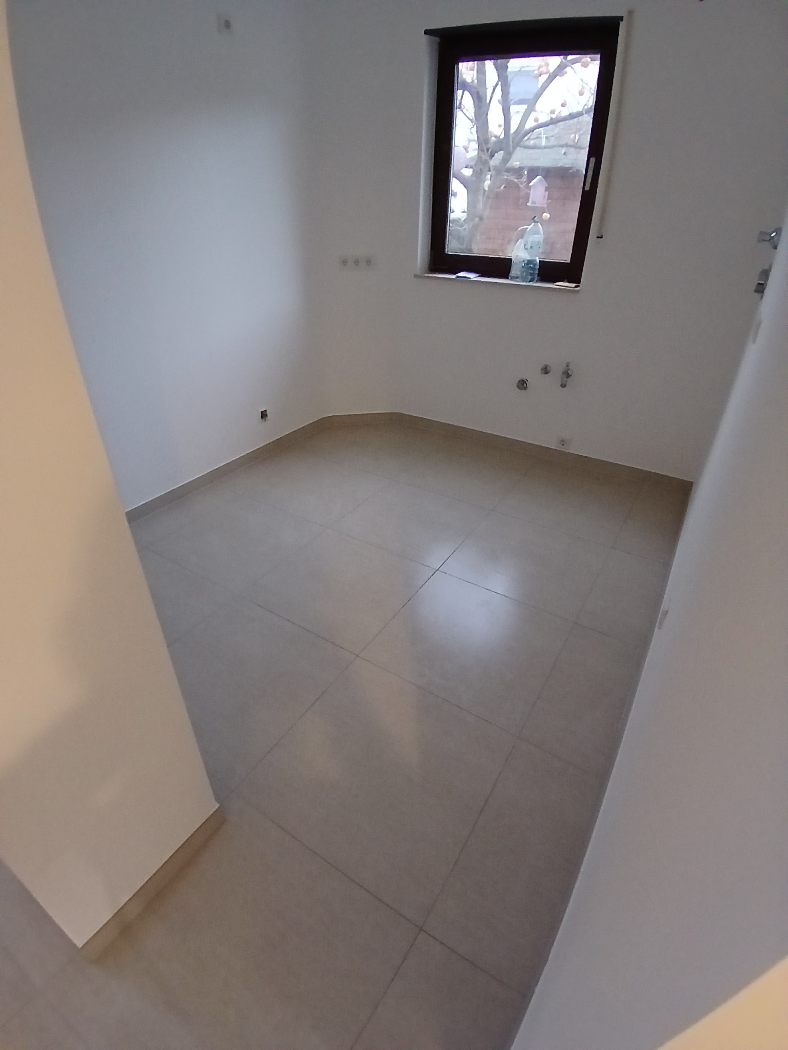 Puste pomieszczenie z beżowymi płytkami podłogowymi, świeżo pomalowanymi białymi ścianami i oknem z widokiem na drzewo z karmnikiem dla ptaków, butelka wody na parapecie.