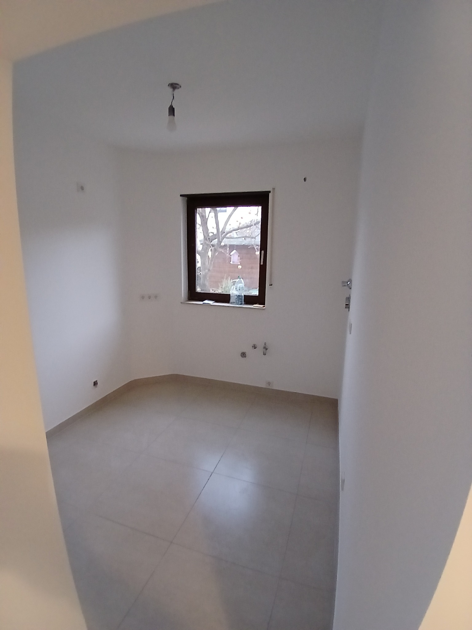 Puste pomieszczenie po remoncie z szarymi płytkami podłogowymi, świeżo pomalowanymi białymi ścianami, oknem z widokiem na podwórze i wiszącą żarówką bez klosza.