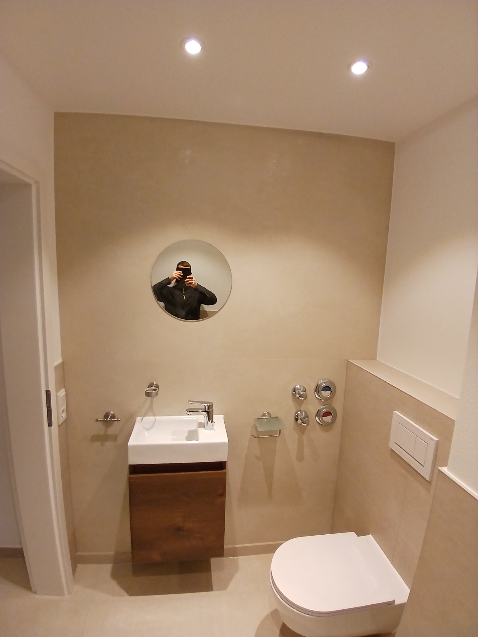 Nowoczesna łazienka z beżowymi ścianami, wiszącą szafką z umywalką z drewna, toaletą i okrągłym lustrem, w którym odbija się osoba robiąca zdjęcie.