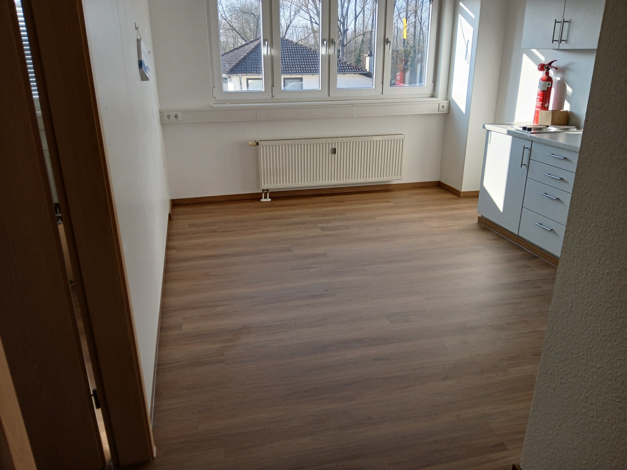 Puste, jasne pomieszczenie z drewnianą podłogą, białymi ścianami, grzejnikiem pod oknem i aneksem kuchennym z gaśnicą, widoczne z perspektywy otwartych drzwi.