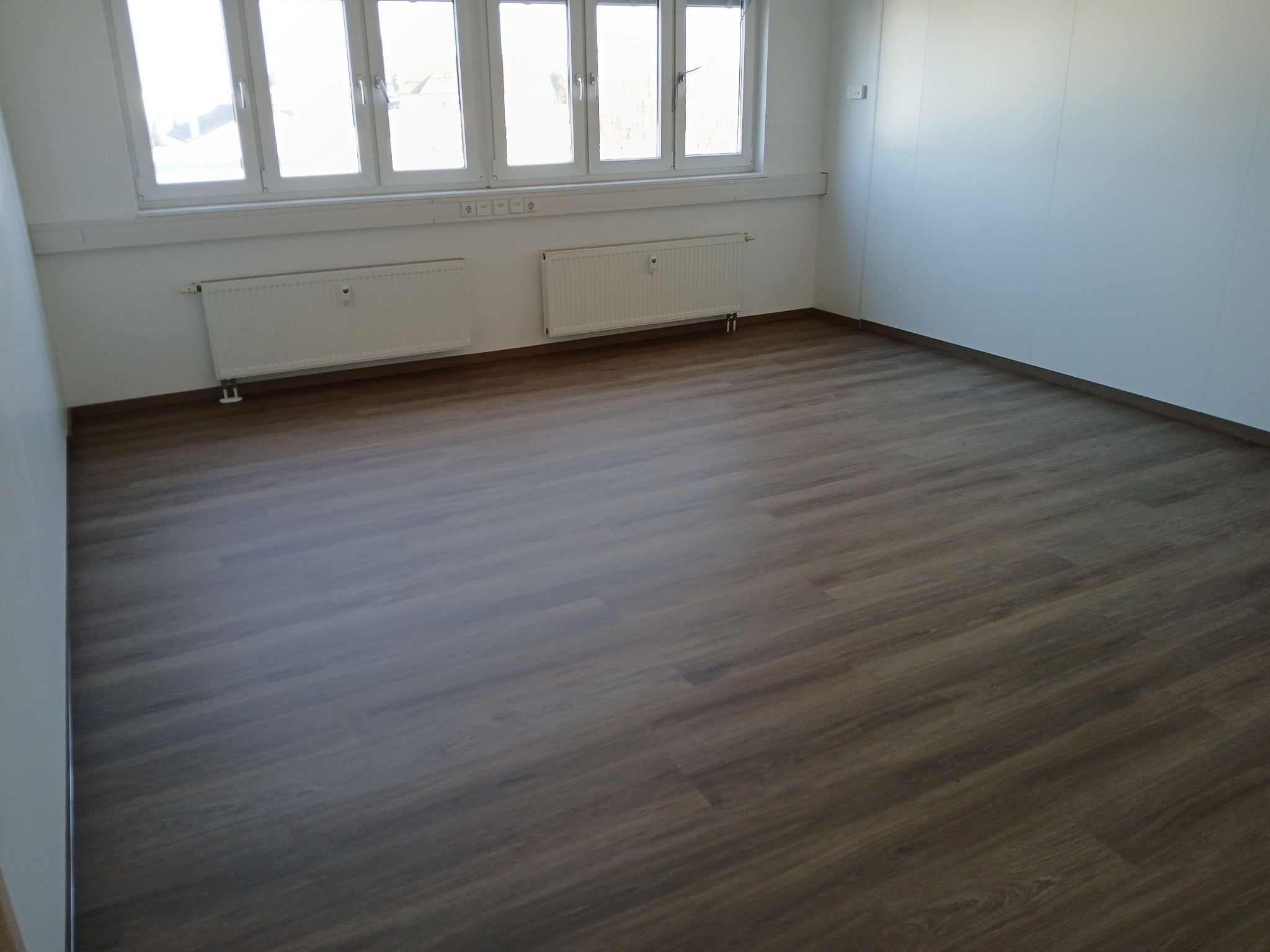 Wykończone wnętrze pokoju z panelami podłogowymi w odcieniach brązu, białymi ścianami i grzejnikami pod oknami.