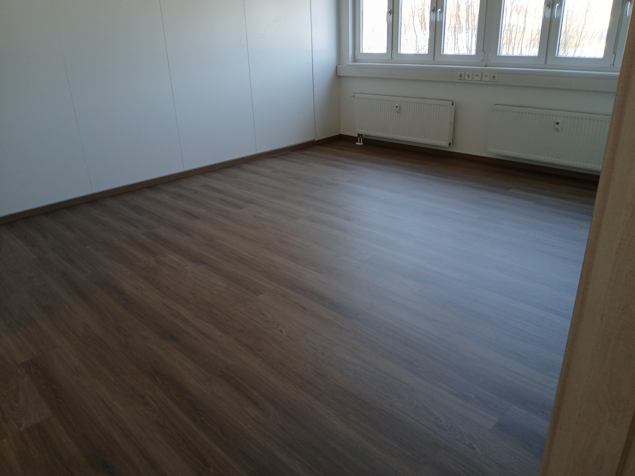 Ułożone panele podłogowe w pustym, jasnym pokoju z białymi ścianami i grzejnikami pod oknami.