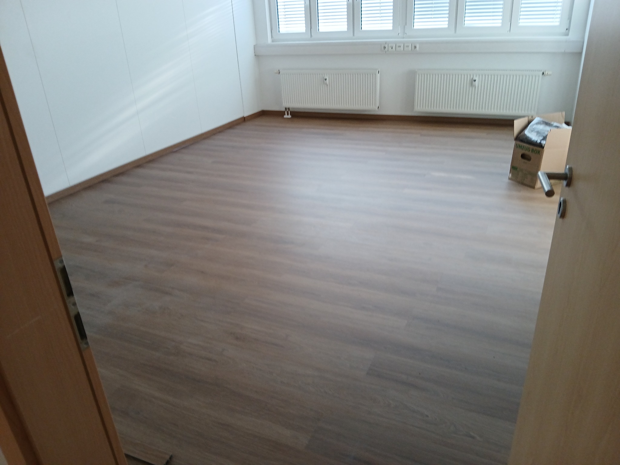 Wnętrze pokoju z ułożonymi panelami podłogowymi o wzorze drewna, widoczne białe ściany, grzejniki pod oknami i kartonowe pudło w rogu, ujęcie z perspektywy otwartych drzwi.