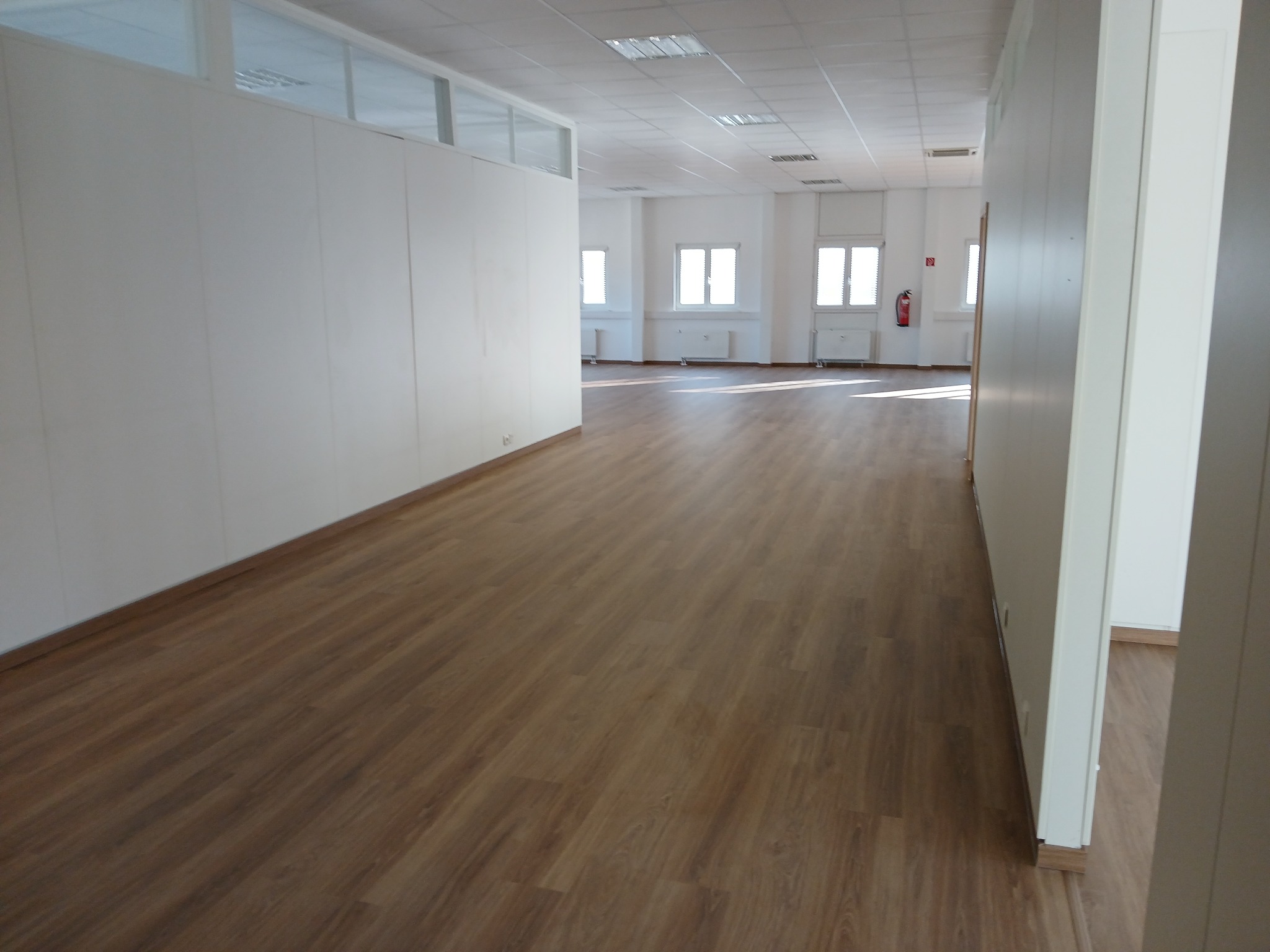 Długi korytarz biurowy z panelami podłogowymi w odcieniach brązu, białe ściany działowe i sufit z oświetleniem rastrowym, widoczne okna z grzejnikami.