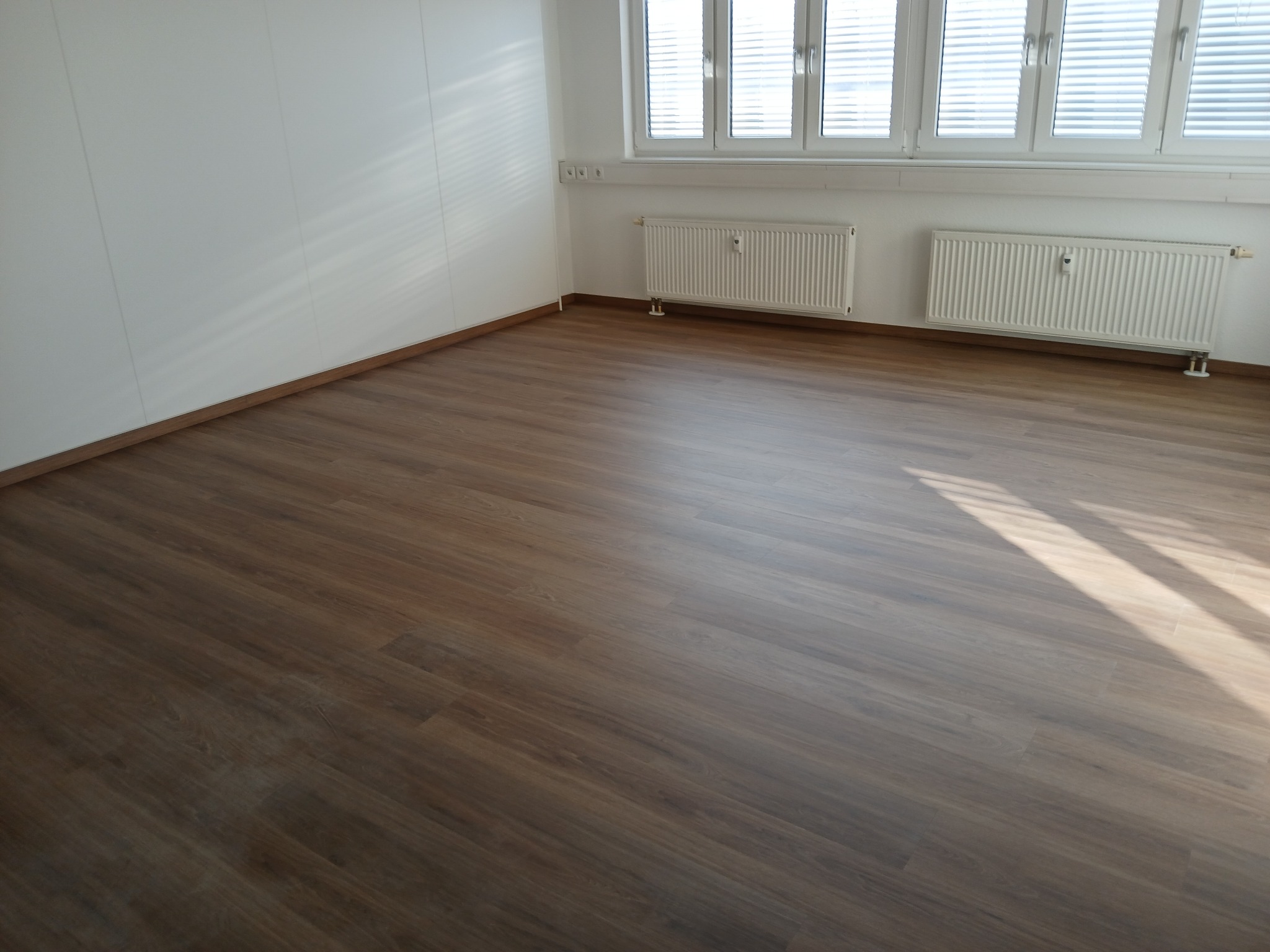 Wnętrze pokoju z ułożonymi panelami podłogowymi o ciemnym odcieniu drewna, widoczne dwa białe grzejniki pod oknem z żaluzjami.