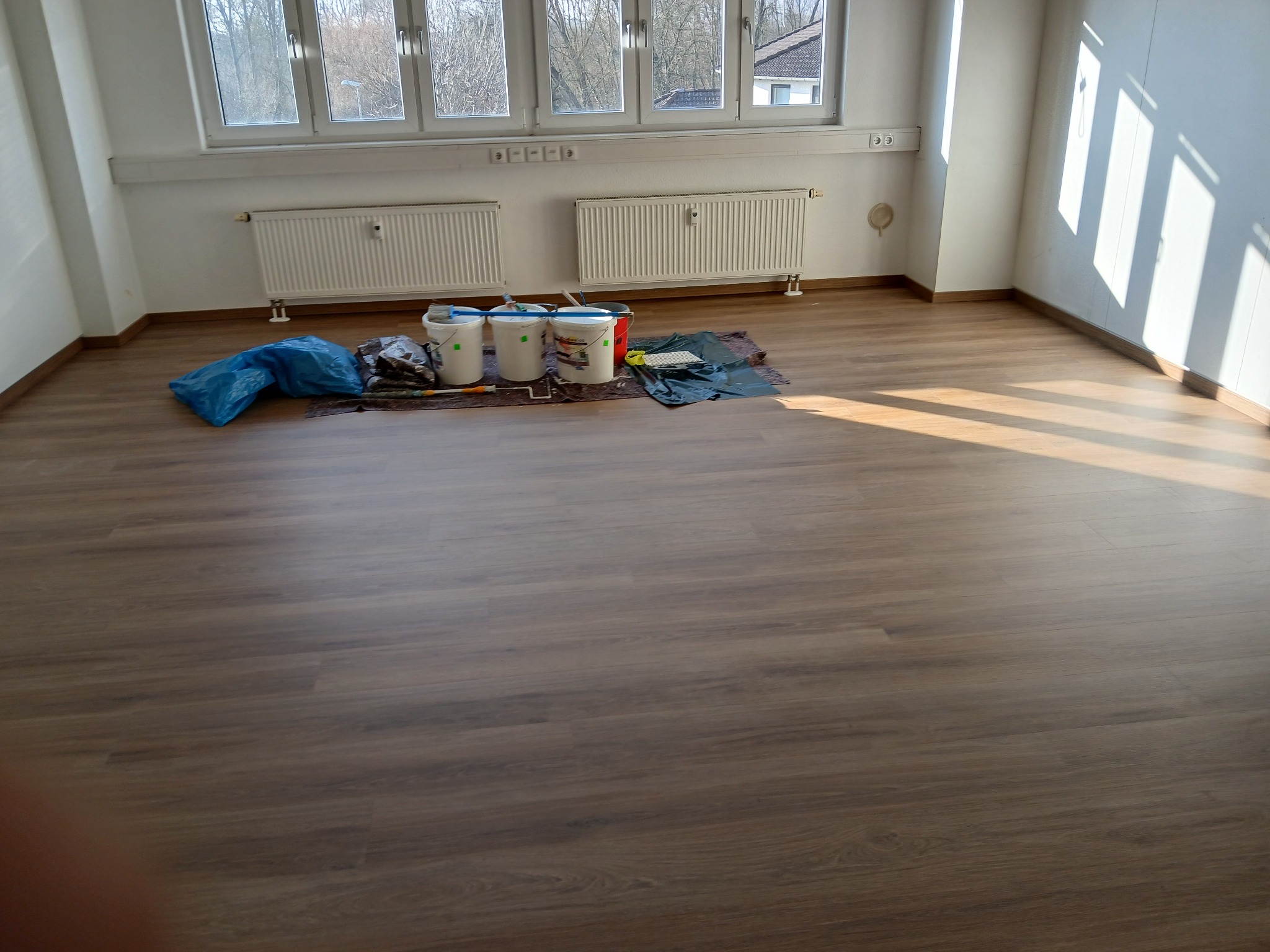 Przygotowanie do prac wykończeniowych w pustym pokoju z drewnianą podłogą: wiadra z farbą, folia ochronna i narzędzia malarskie zgromadzone pod oknami.