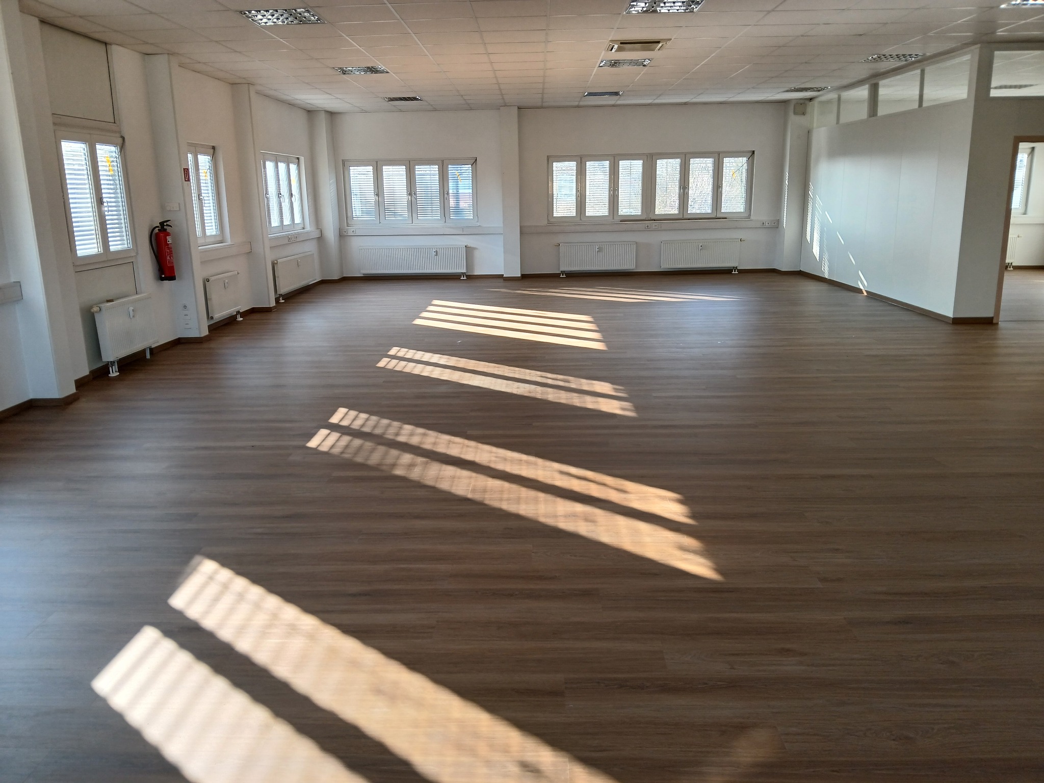 Wnętrze przestronnego, pustego pomieszczenia z drewnianą podłogą, białymi ścianami, grzejnikami pod oknami i wpadającymi promieniami słonecznymi tworzącymi geometryczne wzory na podłodze.