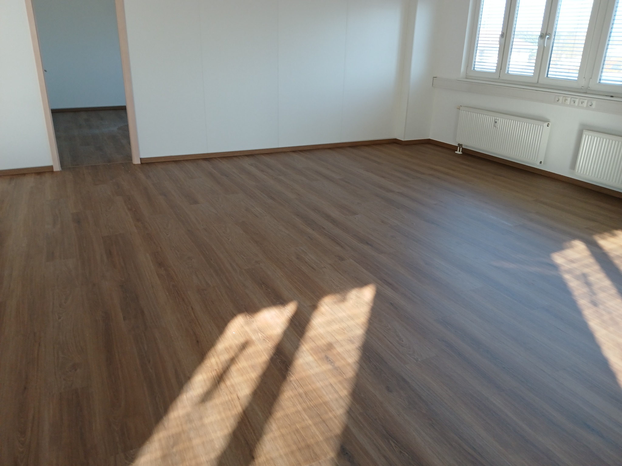 Jasne wnętrze z nowo ułożonymi panelami podłogowymi o wzorze drewna, widoczne promienie słoneczne wpadające przez okno.