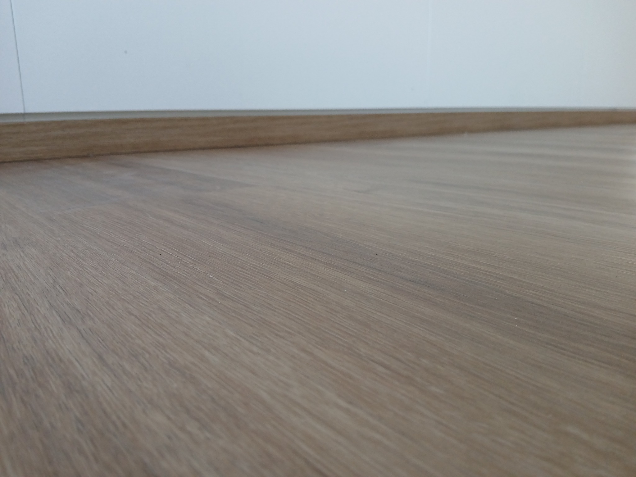 Jasnobrązowe panele podłogowe ułożone wzdłuż ściany z zamontowaną listwą przypodłogową w kolorze drewna.