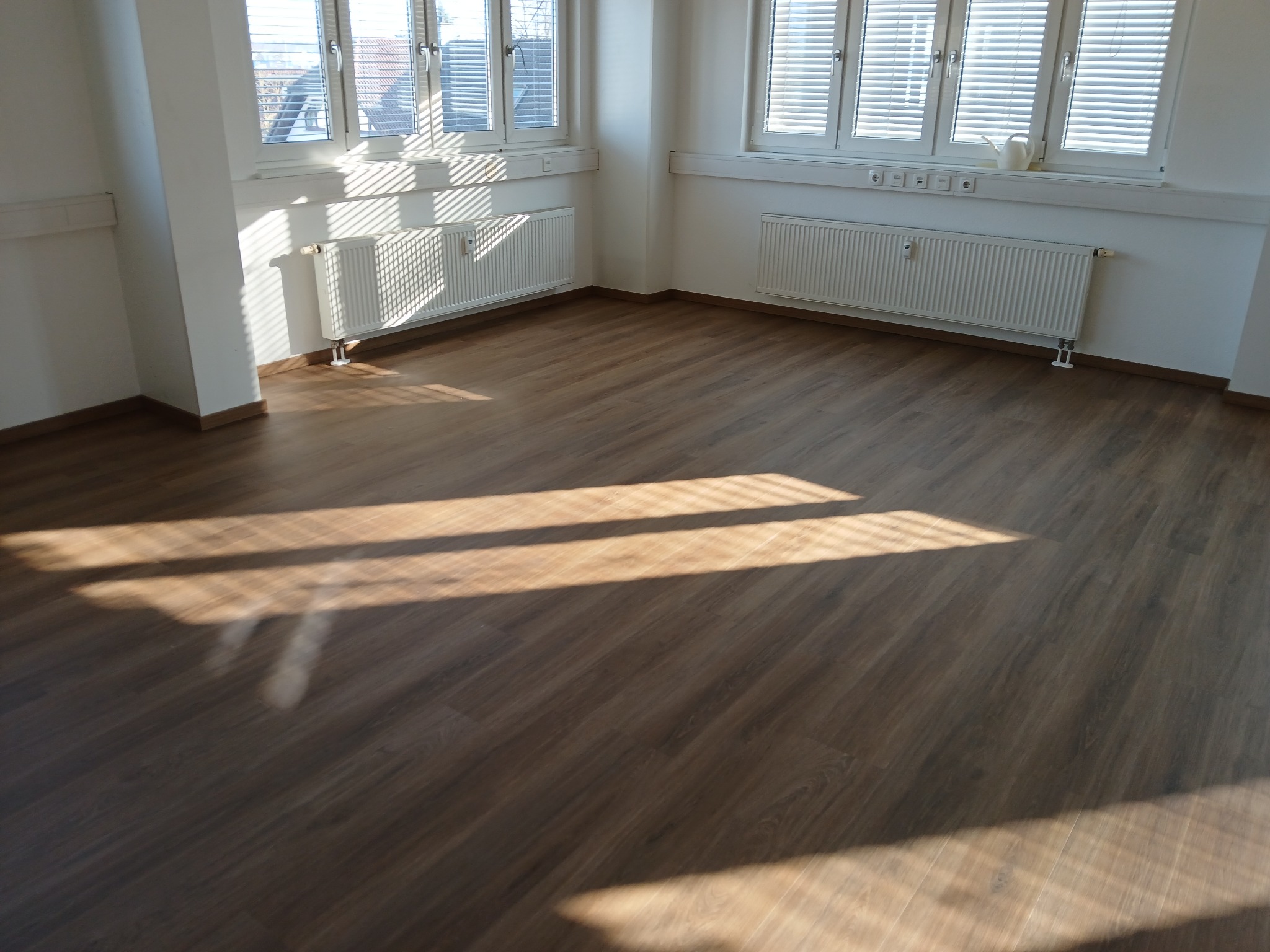 Puste, jasne pomieszczenie z brązową podłogą z paneli, białymi ścianami, grzejnikami i oknami z żaluzjami, na podłodze widoczne promienie słoneczne.