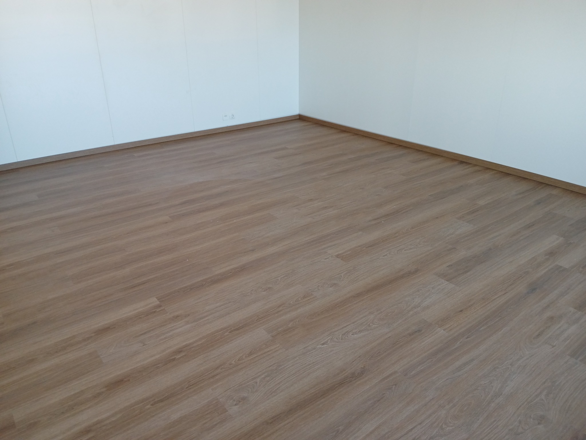 Jasne panele podłogowe imitujące drewno, ułożone w rogu pomieszczenia z białymi ścianami i listwami przypodłogowymi.