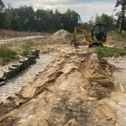 Żółta mini koparka wyrównuje teren pod budowę chodnika z szarej kostki brukowej, widoczne ślady opon i hałdy piasku, w tle las.
